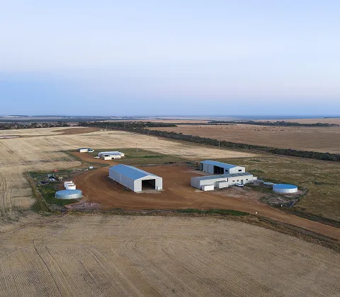 Aerial view of a rural area featuring multiple buildings including warehouses and storage tanks on a dirt path, surrounded by expansive fields of golden crops and sparse trees under a clear sky.