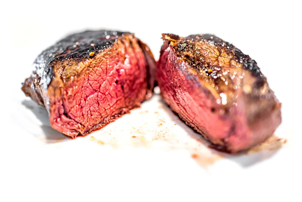 Two pieces of sliced beef tenderloin laid on a white surface. The outer layer shows a charred surface, while the inside displays a medium-rare pink center.